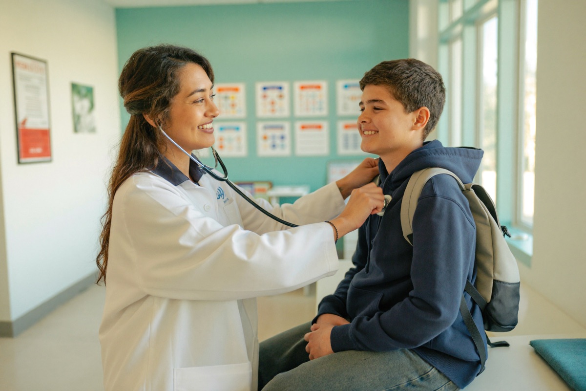 A doctor listens to a smiling teenage student's chest with a stethoscope during a back-to-school checkup