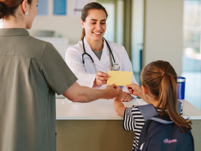 A parent and her daughter hand a yellow immunization card to clinic staff at the reception desk